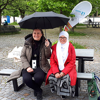 zwei Frauen sitzen lachend auf einer Bank uner einem Regenschirm