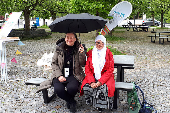 zwei Frauen sitzen lachend auf einer Bank uner einem Regenschirm