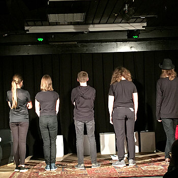 Fünf schwarz gekleidete Jugendliche vor schwarzem Hintergrund aud der Bühne. Sie stehen mit dem Rücken zum Publikum