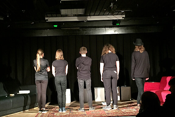Fünf schwarz gekleidete Jugendliche vor schwarzem Hintergrund aud der Bühne. Sie stehen mit dem Rücken zum Publikum