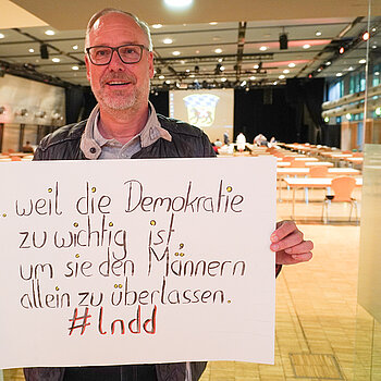 Verwaltungsdirektor Gerhard Koch hält ein Schild mit der Aufschrift "weil die Demokratie zu wichtig ist, um sie den Männern allein zu überlassen. "