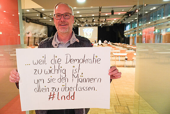 Verwaltungsdirektor Gerhard Koch hält ein Schild mit der Aufschrift "weil die Demokratie zu wichtig ist, um sie den Männern allein zu überlassen. "