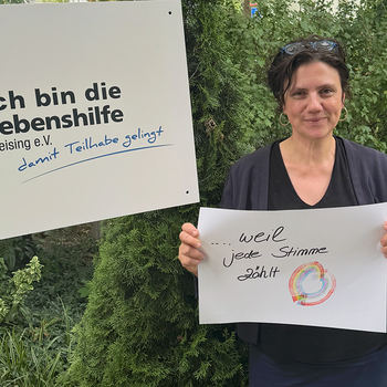 Nadja Stahlberger feiert die Demokratie, weil jede Stimme zählt. Foto: Lebenshilfe Eine Frau mit Brille im Haar steht im Grünen und hält ein Schild, daneben hält jemand ein Schild "Ich bin die Lebenshilfe Freising e.V."