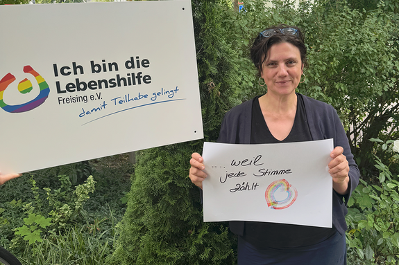 Nadja Stahlberger feiert die Demokratie, weil jede Stimme zählt. Foto: Lebenshilfe Eine Frau mit Brille im Haar steht im Grünen und hält ein Schild, daneben hält jemand ein Schild "Ich bin die Lebenshilfe Freising e.V."