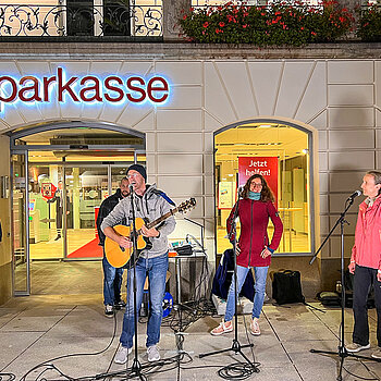 Vor dem beleuchteten Eingang der Sparkasse stehen ein Mann mit Gitarre und zwei Sängerinnen sowie zwei Zuhörer.