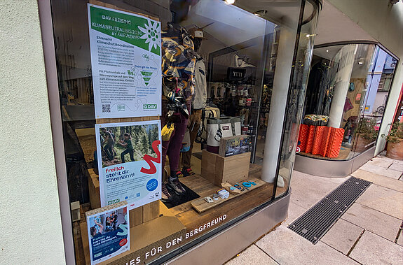Das DAV-Fenster im Freiluftwerk warb für Engagement für Klimaschutz. Foto: Stadt Freising Neben Schaufensterpuppen in Bergbekleidung hängen 2 Plakate: "Klimaneutral bis 2030" und Menschen, die Wege instand setzen.