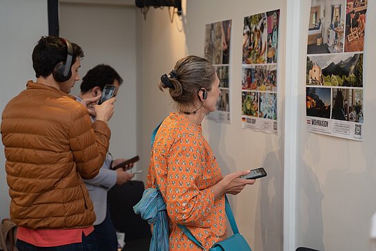 Blick in die Ausstellung "Solution Exhibition". Foto: Snowball Effekt Menschen stehen mit Kopfhörern vor Plakaten und betrachten sie.