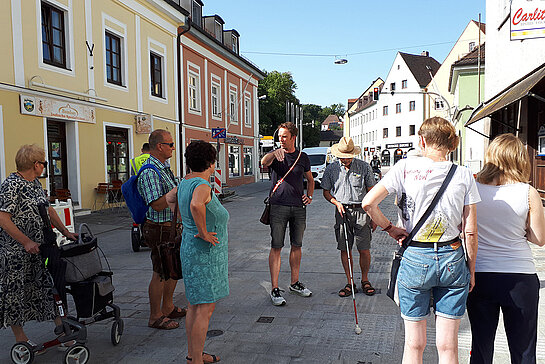 mehrer Menschen, teilweise mit Rollator und Langstock stehen auf der Straße und reden miteinander