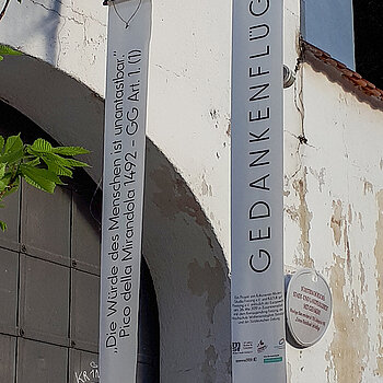 Tor und Fassade des Alten Gefängnisses. Vor dem braunen Tor hängen zwei lange Fahnen: "Gedankenflüge" und "Die Würde des Menschen ist unantastbar"
