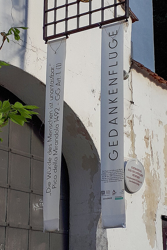 Tor und Fassade des Alten Gefängnisses. Vor dem braunen Tor hängen zwei lange Fahnen: "Gedankenflüge" und "Die Würde des Menschen ist unantastbar"