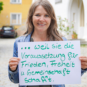 Claudia Bauer (Kreisbildungswerk Freising) feiert die Demokratie, weil sie die Voraussetzung für Frieden, Freiheit und Gemeinschaft schafft. Foto: Stadt Freising Eine Frau mit langen Haaren steht im Freien und hält ein Schild. Im Hintergrund Kirchenportal, Baum und Auto.