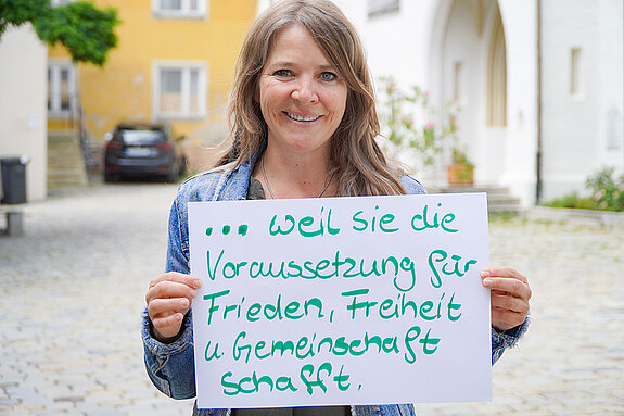 Claudia Bauer (Kreisbildungswerk Freising) feiert die Demokratie, weil sie die Voraussetzung für Frieden, Freiheit und Gemeinschaft schafft. Foto: Stadt Freising Eine Frau mit langen Haaren steht im Freien und hält ein Schild. Im Hintergrund Kirchenportal, Baum und Auto.