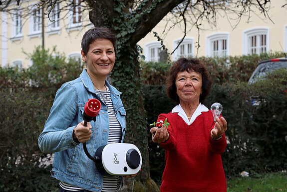 zwei Frauen vor einer grünen Hecke, eine mit Glühbirne und eine Pflanze. Eine trägt eine Wallbox.