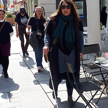 Eine Frau mit Sonnenbrille und Langstock stößt auf einen Stuhl 