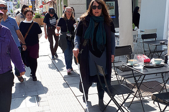 Eine Frau mit Sonnenbrille und Langstock stößt auf einen Stuhl 