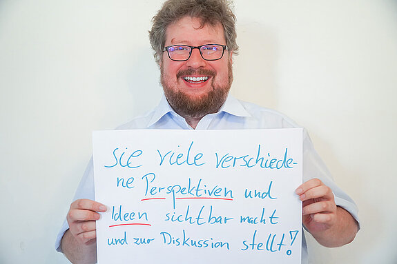 Stadtratsmitglied Dr. Maximilian Breu. Foto: Stadt Freising Ein Mann in einem weißen Hemd zeigt ein Schild mit der Aufschrift "sie viele verschiedene Perspektiven und deen sichtbar macht und zur Diskussion stellt!
