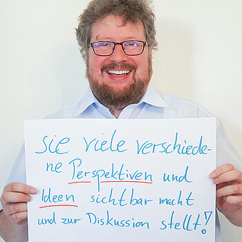 Stadtratsmitglied Dr. Maximilian Breu. Foto: Stadt Freising Ein Mann in einem weißen Hemd zeigt ein Schild mit der Aufschrift "sie viele verschiedene Perspektiven und deen sichtbar macht und zur Diskussion stellt!
