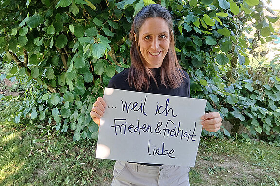 Johanna Weinberger feiert die Demokratie, weil sie Frieden und Freiheit liebt. Foto: Johanna Weinberger Eine Frau mit längeren Haaren und einem dunklen Oberteil steht mit einem Schild vor einer Hecke .