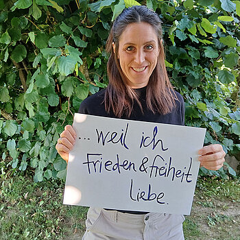 Johanna Weinberger feiert die Demokratie, weil sie Frieden und Freiheit liebt. Foto: Johanna Weinberger Eine Frau mit längeren Haaren und einem dunklen Oberteil steht mit einem Schild vor einer Hecke .