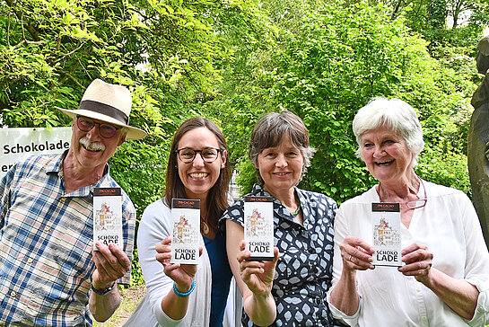 drei Frauen und ein Mann halten lachend Schokoladentafeln vor sich. Im Hintergrund Büsche