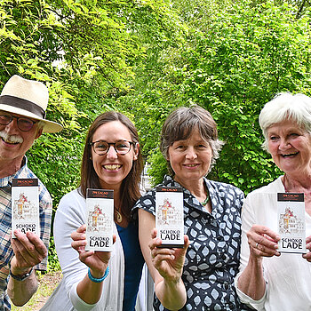 drei Frauen und ein Mann halten lachend Schokoladentafeln vor sich. Im Hintergrund Büsche