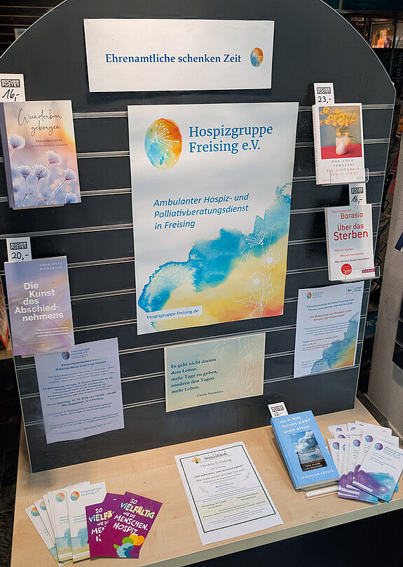 Ehrenamtliche schenken Zeit - bei der Hospizgruppe im Fenster bei Pustet. Foto: Stadt Freising ein Plakat der Hospizgruppe in hellblau und orange-Tönen umgeben von mehreren Büchern zu Tod und Trauer.
