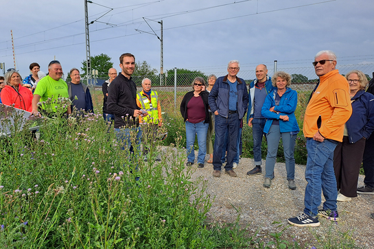 Simon Prechsl von den Freisinger Stadtwerken erklärt die Funktion der PV-Anlage. Foto: Sepp Beck eine Gruppe Menschen in bunter Kleidung steht auf einem Weg. Im Hintergrund Zaun und Bahnstrecke, vorne Wiese und ein Solarmodul.
