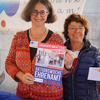 zwei Frauen halten lachend ein Plakat und einen Flyer in die Kamera