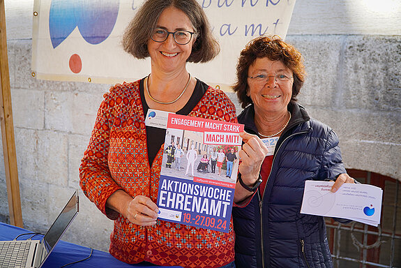 zwei Frauen halten lachend ein Plakat und einen Flyer in die Kamera
