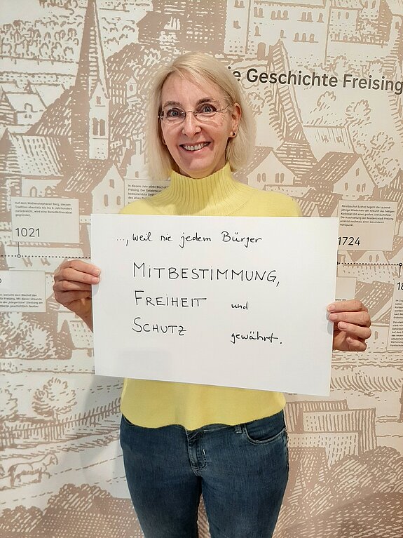 Eva Willberg feiert die Demokratie, weil sie jedem Bürger Mitbestimmung, Freiheit und Schutz gewährt. Foto: Stadt Freising Eine blonde Frau mit Brille und im gelben Pullover steht vor einer Wand "Geschichte Freisings" und hält ein Schild in den Händen.