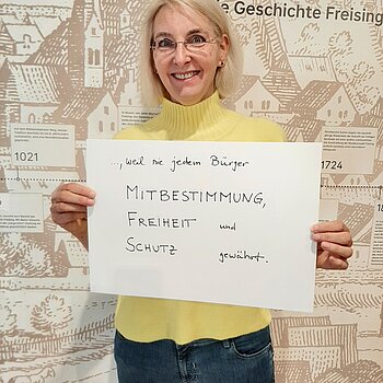 Eva Willberg feiert die Demokratie, weil sie jedem Bürger Mitbestimmung, Freiheit und Schutz gewährt. Foto: Stadt Freising Eine blonde Frau mit Brille und im gelben Pullover steht vor einer Wand "Geschichte Freisings" und hält ein Schild in den Händen.