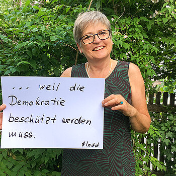 Christel Orthen-Schmuker, IT-Dozentin steht vor einer Hecke und hält ein Schild mit der Aufschrift:  " weil die Demokratie beschützt werden muss."