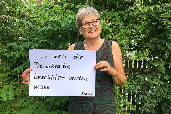 Christel Orthen-Schmuker, IT-Dozentin steht vor einer Hecke und hält ein Schild mit der Aufschrift:  " weil die Demokratie beschützt werden muss."