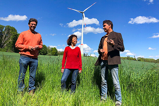 Zwei Männer und eine Frau in einem Feld. Im Hintergrund ein Windrad.