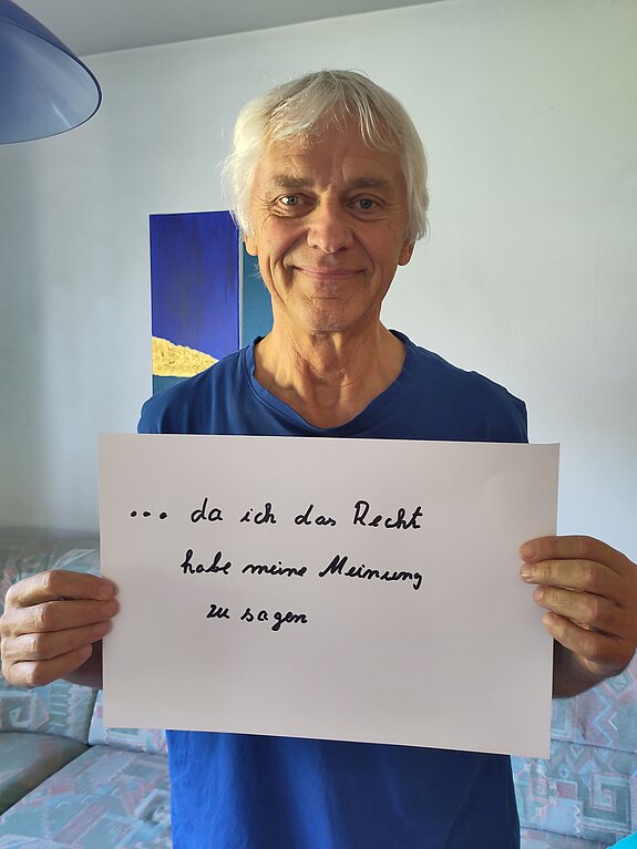 Ansgar Wübbels (Agenda21) feiert die Demokratie, weil er da das Recht hat, seine Meinung zu sagen. Foto: Wübbels Ein grauhaariger Mann im blauen Shirt steht in einem Raum und hält ein Schild