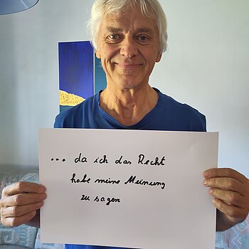 Ansgar Wübbels (Agenda21) feiert die Demokratie, weil er da das Recht hat, seine Meinung zu sagen. Foto: Wübbels Ein grauhaariger Mann im blauen Shirt steht in einem Raum und hält ein Schild