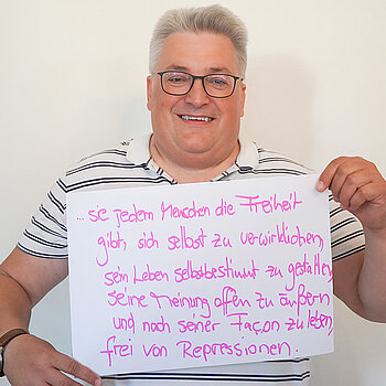 Stadtratsmitglied Werner Habermeyer. Foto: Stadt Freising Ein Mann mit Brille und einem grau weiß gestreiften T-Shirt zeigt ein Schild mit der Aufschrift "sie jedem Menschen die Freiheit gibt, sich selbst zu verwirklichen, sein Leben selbstbestimmt zu gestalten, seine Meinung offen zu äußern und nach seiner Facon zu leben, frei von Repressionen