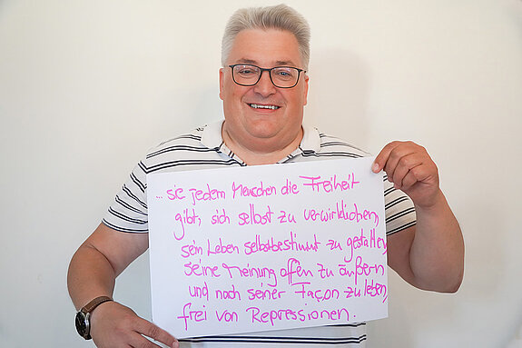 Stadtratsmitglied Werner Habermeyer. Foto: Stadt Freising Ein Mann mit Brille und einem grau weiß gestreiften T-Shirt zeigt ein Schild mit der Aufschrift "sie jedem Menschen die Freiheit gibt, sich selbst zu verwirklichen, sein Leben selbstbestimmt zu gestalten, seine Meinung offen zu äußern und nach seiner Facon zu leben, frei von Repressionen