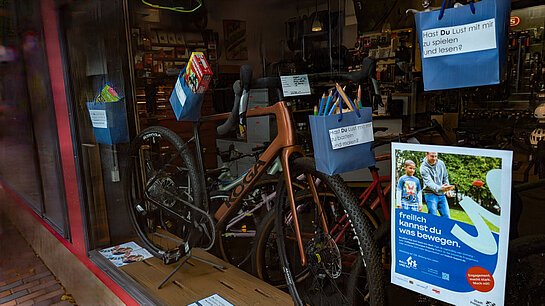 Balu und Du bei Radl Ruhland zeigte die Vielfalt der Möglichkeiten, was man unternehmen kann mit Mogli. Foto: Stadt Freising Neben einem Fahrrad in einem Fenster ist ein Plakat angeklebt und mehrere blaue Papiertüten hängen mit Spiel und Bastelmaterial daneben.