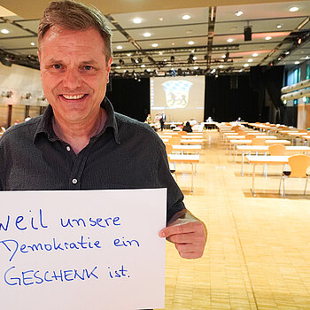 Stadtrat Peter Warlimont hält ein Schild mit der Aufschrift: "weil unsere Demokratie ein Geschenk ist." 