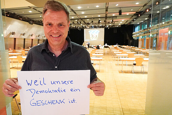 Stadtrat Peter Warlimont hält ein Schild mit der Aufschrift: "weil unsere Demokratie ein Geschenk ist." 