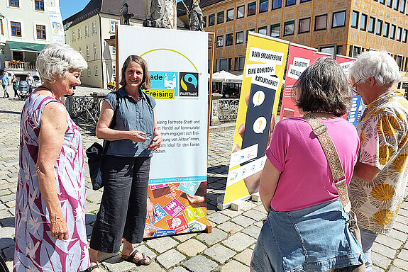 zwei Frauen stehen vor großen Bannern einer Ausstellung und sprechen mit einem Paar, das von hinten zu sehen ist.