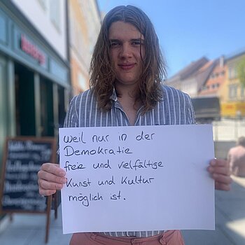 Jonas Günther (Musiker und Organisator Jung & Laut) feiert die Demokratie, weil nur in der Demokratie freie und vielfältige Kultur möglich ist. Foto: privat Ein junger Mann mit langen Haaren steht in der Freisinger Innenstadt und hält ein Schild.