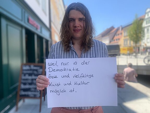 Jonas Günther (Musiker und Organisator Jung & Laut) feiert die Demokratie, weil nur in der Demokratie freie und vielfältige Kultur möglich ist. Foto: privat Ein junger Mann mit langen Haaren steht in der Freisinger Innenstadt und hält ein Schild.