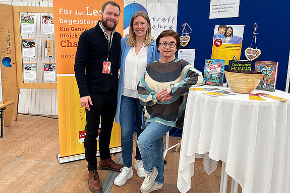 Zwei Frauen und ein Mann stehen an einem Stehtischmit Büchern. Dahinter ein orangefarbenes Rollup mit 