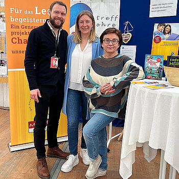 Zwei Frauen und ein Mann stehen an einem Stehtischmit Büchern. Dahinter ein orangefarbenes Rollup mit 