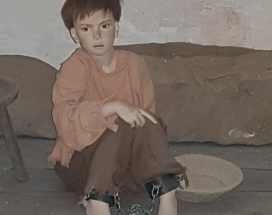 Puppe eines Buben von vielleicht 10 Jahren in schlechter Kleidung am Boden im Gefängnis sitzend.