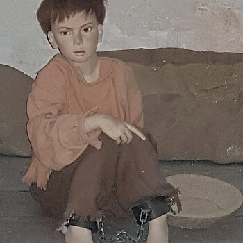 Puppe eines Buben von vielleicht 10 Jahren in schlechter Kleidung am Boden im Gefängnis sitzend.