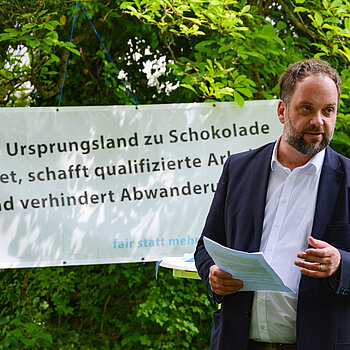 Ein Mann mit Papieren in der Hand vor einem Banner und Büschen.