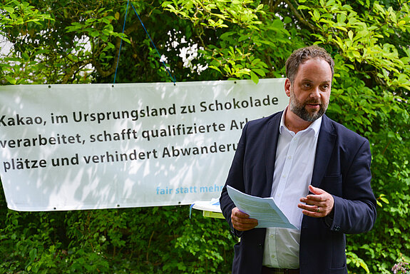 Ein Mann mit Papieren in der Hand vor einem Banner und Büschen.